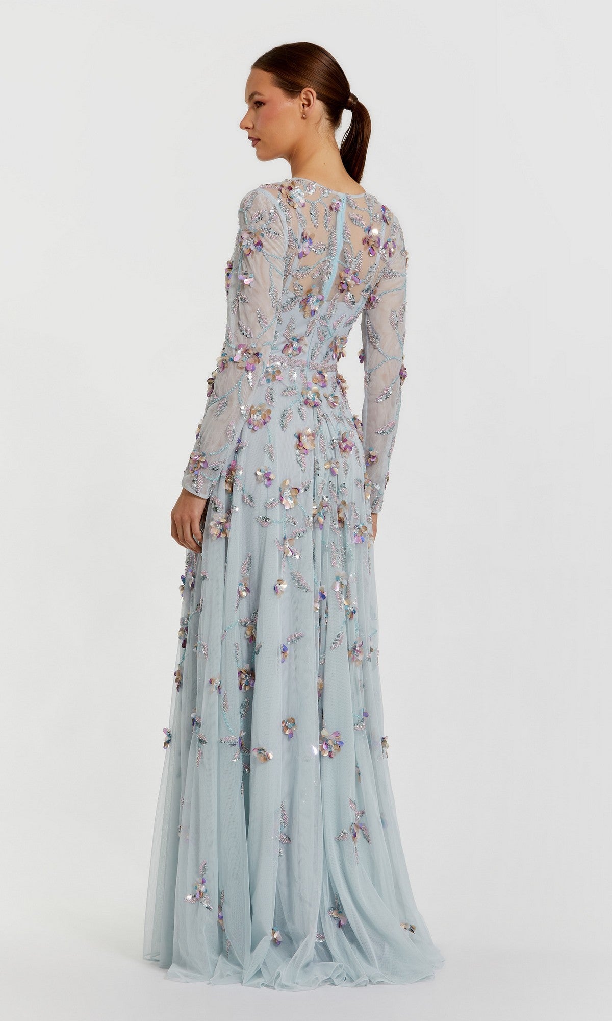 A woman stands in profile wearing the Mac Duggal 6183 Long Sleeve Beaded Formal Gown, a light blue floor-length dress adorned with floral appliqué detailing.